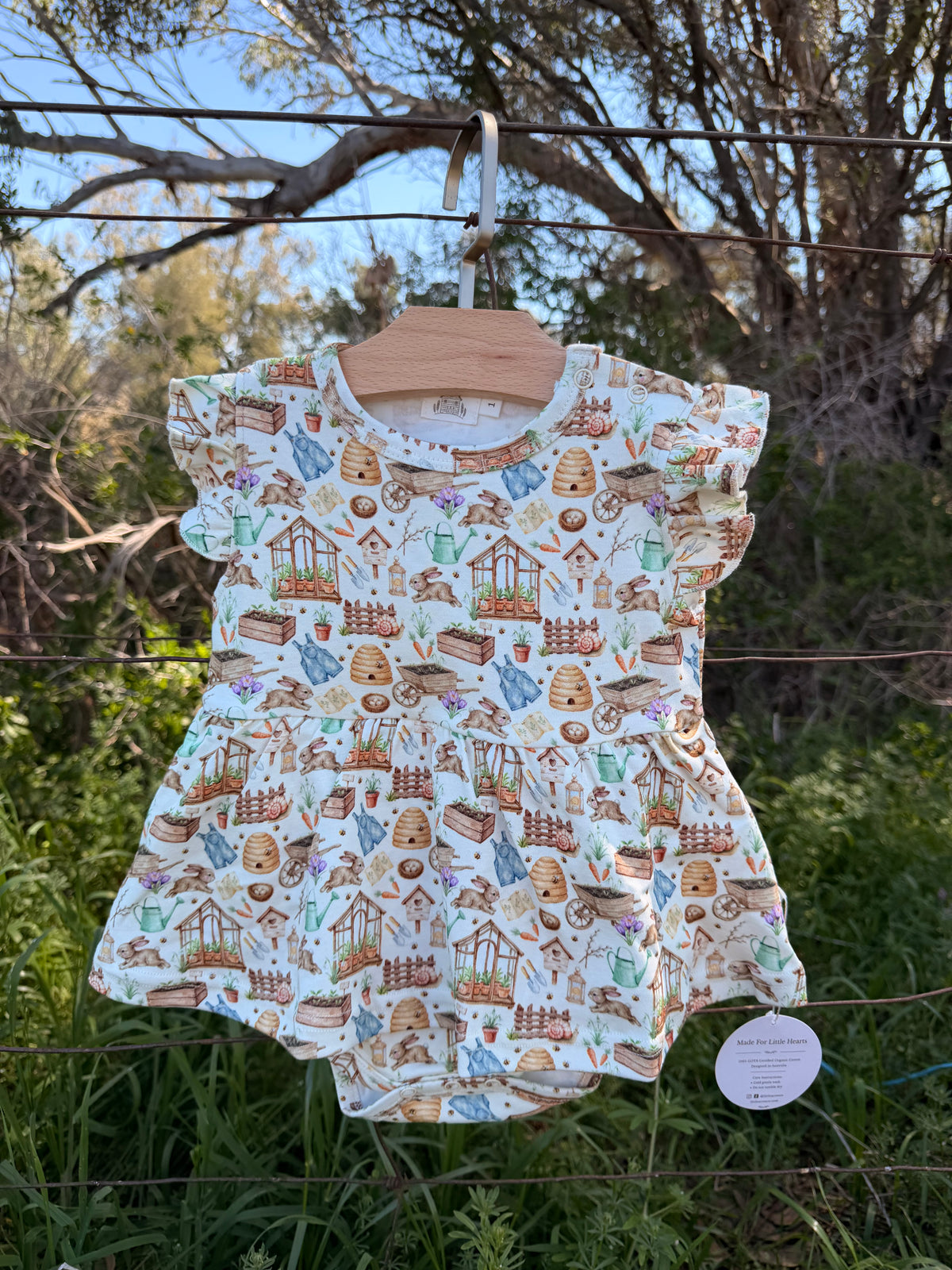 A baby romper hanging on a wooden hanger outdoors on a wire fence, featuring a colorful pattern of garden elements like beehives, carrots, and flowers on a light blue background.