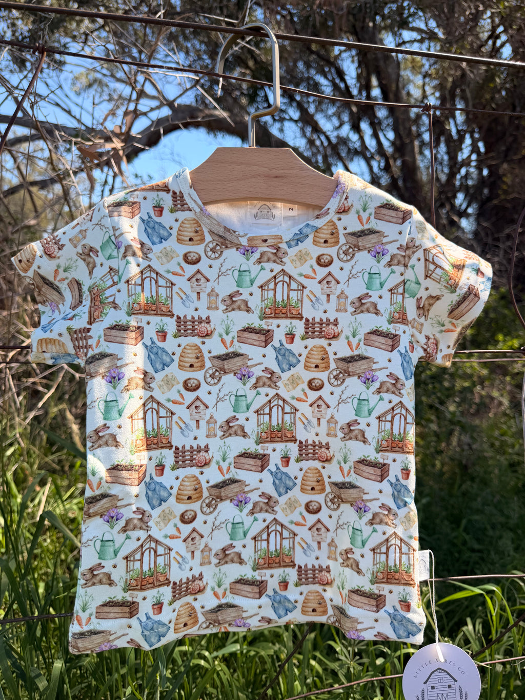 A short-sleeve t-shirt with a garden-themed all-over print, hanging on a wooden hanger outdoors on a wire fence. The print features illustrations of gardening tools, pots, and plants on a light cream background.