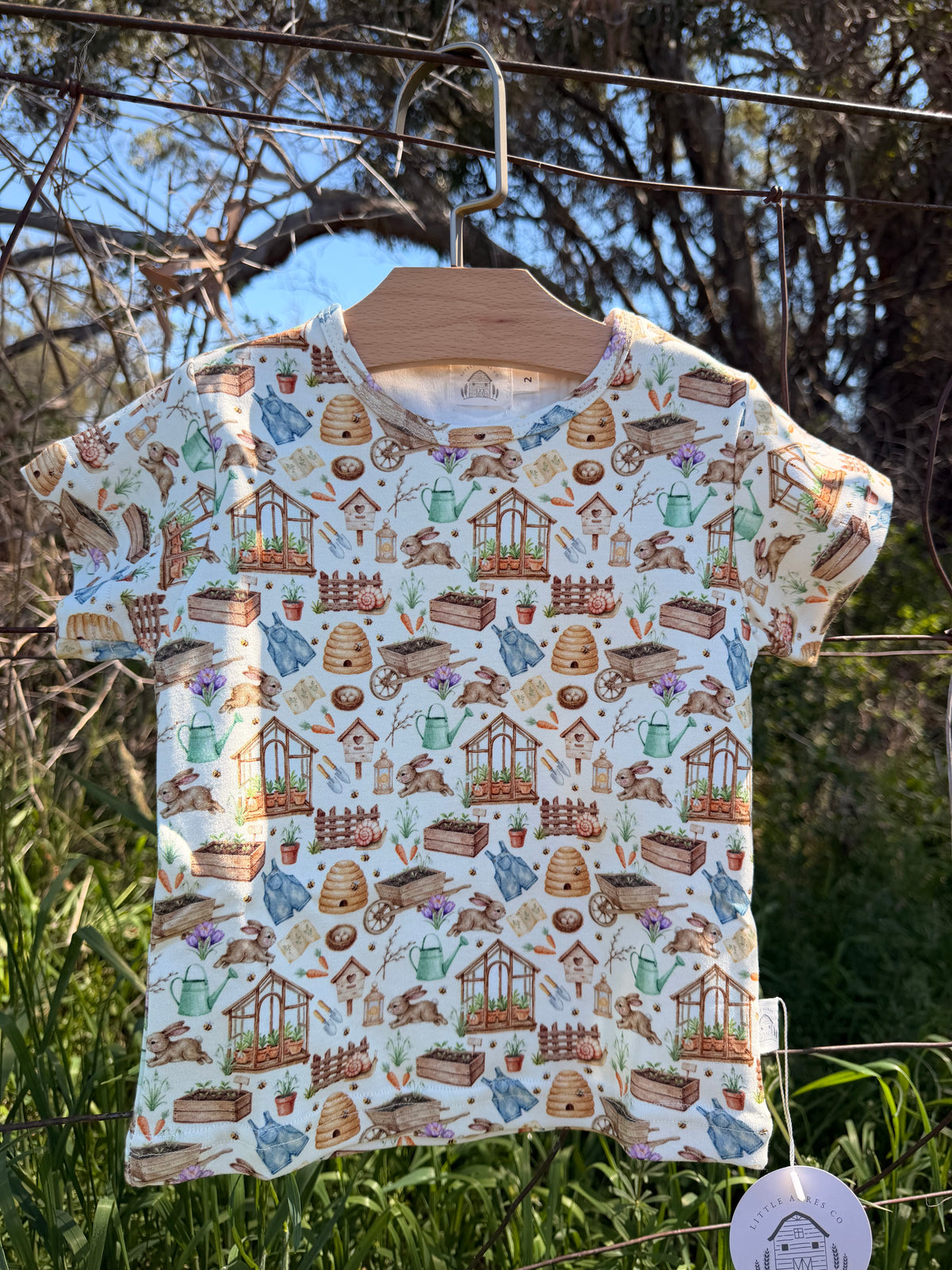 A short-sleeve t-shirt with a garden-themed all-over print, hanging on a wooden hanger outdoors on a wire fence. The print features illustrations of gardening tools, pots, and plants on a light cream background.