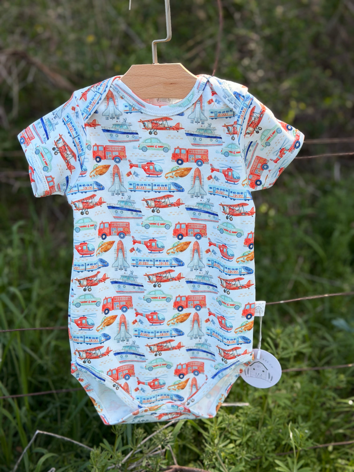 A baby short-sleeve bodysuit with a colorful fire station and vehicle print, hanging on a wooden hanger outdoors. From company little acres co 