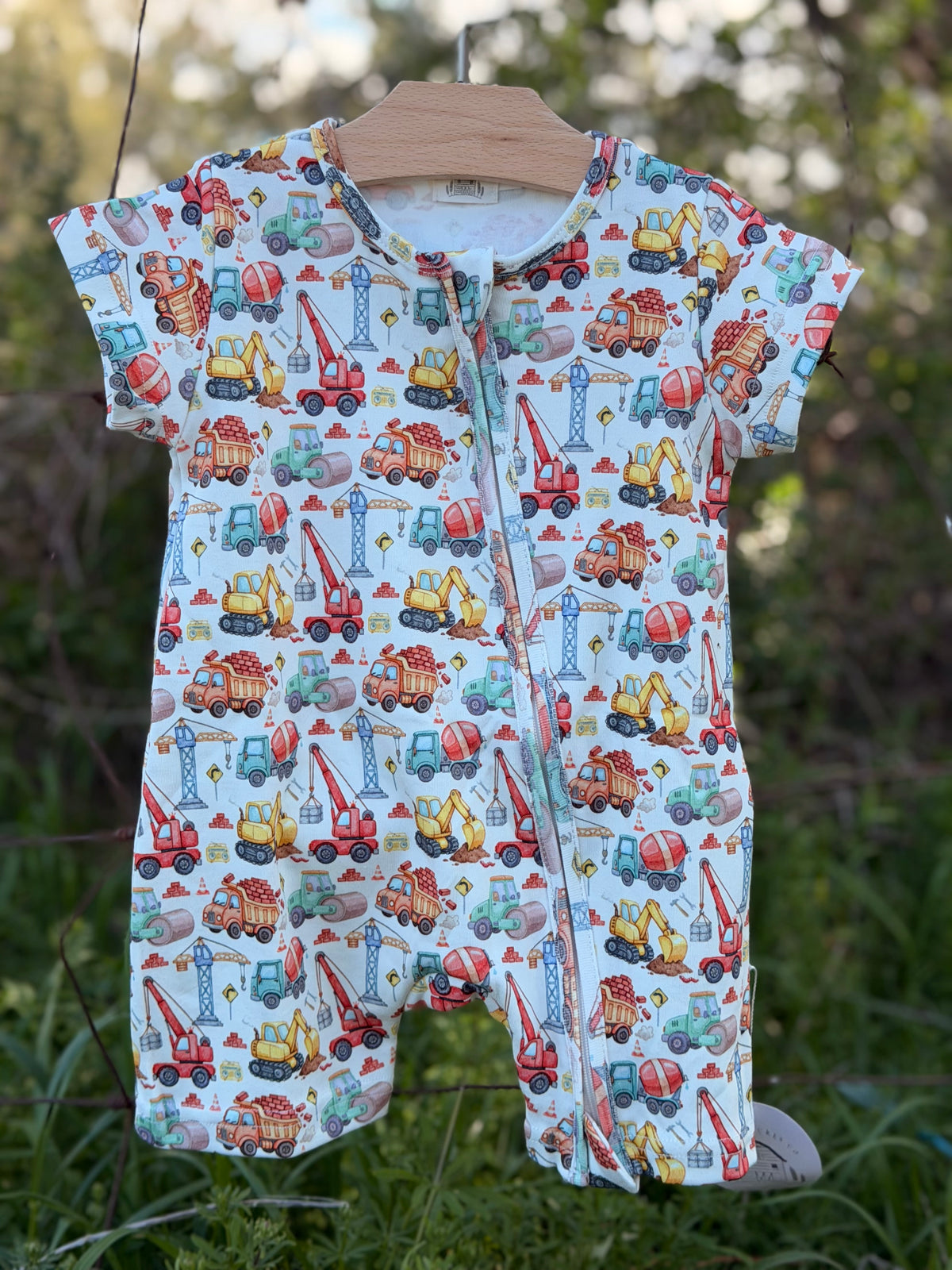 A baby short-sleeve footless romper with a colorful construction vehicle print, hanging on a wooden hanger from company Little Acres Co