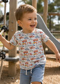 A young boy smiling and laughing on a playground, wearing a little Acres Co T-shirt which has a colorful pattern of airplanes, rockets, and cars on a white background.
