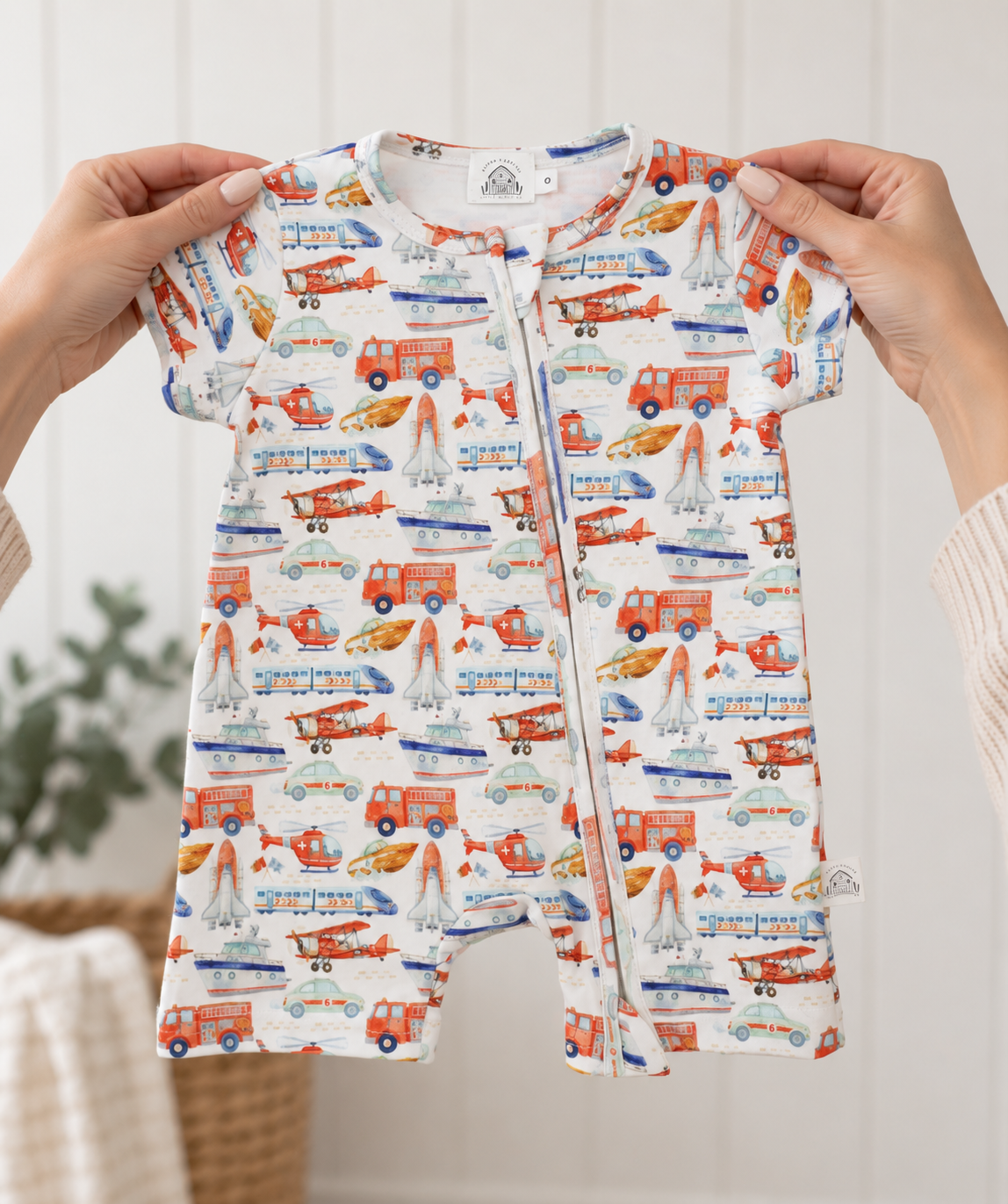 Little Acres Co transport print baby zip-up romper in GOTS organic cotton. The romper has an all-over colorful print of red fire trucks, blue helicopters, orange airplanes, and white sailboats on a cream background.
